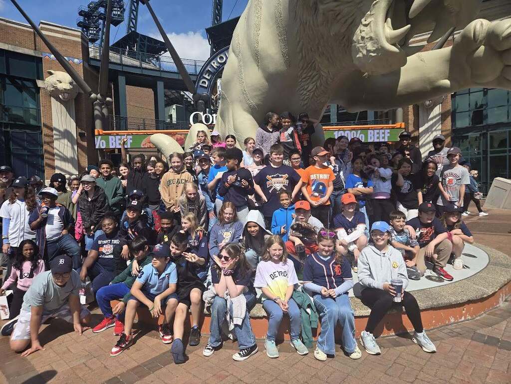Large group of children and adults posing in front of a giant tiger statue at an outdoor plaza on a sunny day.