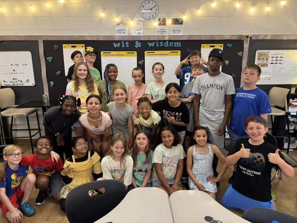 Group of diverse elementary students posing together in a classroom decorated with 'wit & wisdom' posters and string lights.