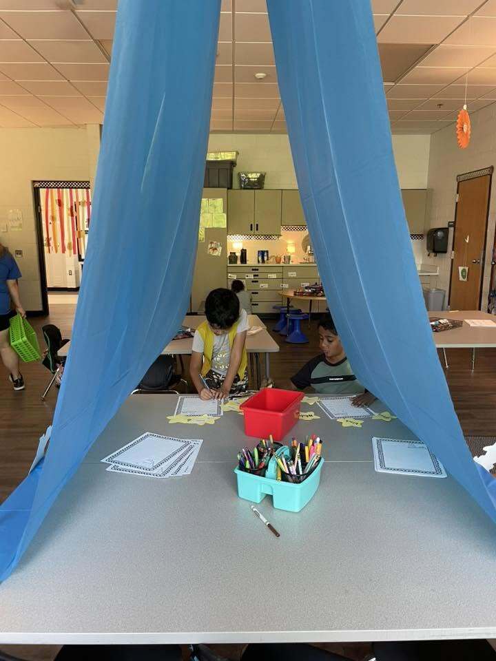 Two children engaged in a classroom activity at a table partially enclosed by blue fabric panels hanging from above.