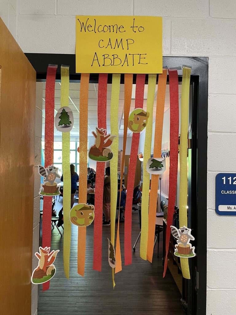 Classroom door decorated with colorful streamers and woodland animal cutouts under a yellow sign reading 'Welcome to CAMP ABBATE'.