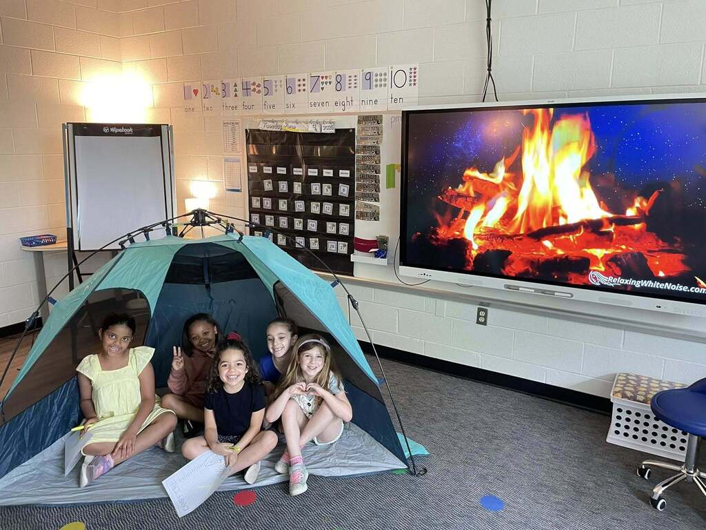 Five children sitting inside a small indoor tent in a classroom with a large screen showing a virtual campfire on the wall.