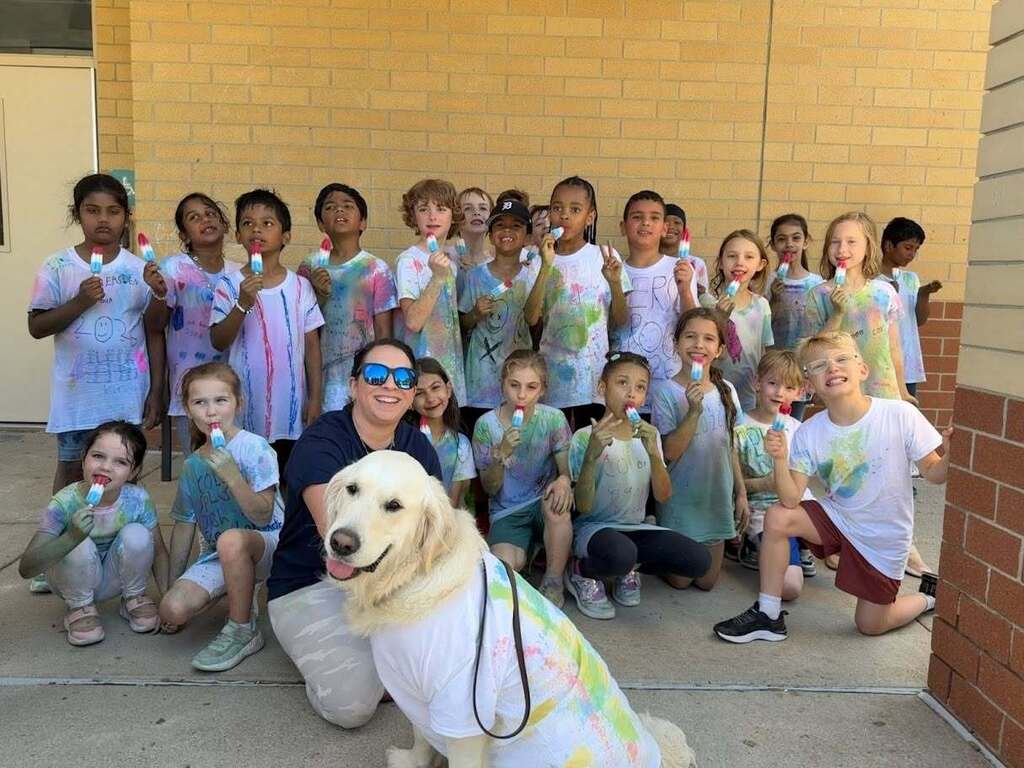 Group of children wearing colorful tie-dye shirts posing outdoors with a large white dog also dressed in a tie-dye shirt.