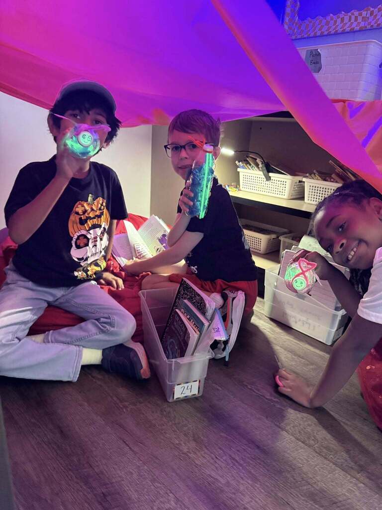 Three children sitting and playing under a colorful fabric fort indoors with storage bins and toys around them.