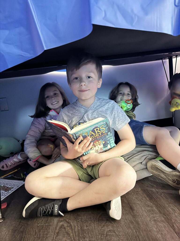 Three children sitting under a blanket fort, one holding and reading a book titled 'Stars'.