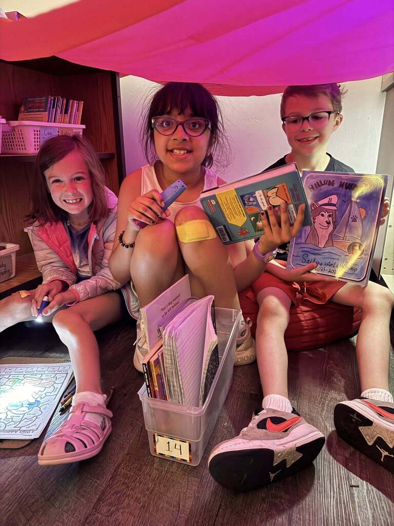 Three children sitting under a pink fabric fort, reading books and using flashlights in a cozy indoor setting.