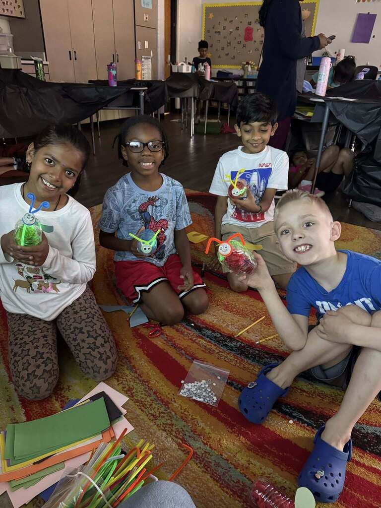 Four children sitting on a colorful rug in a classroom, each holding a decorated plastic bottle craft with straws and beads.