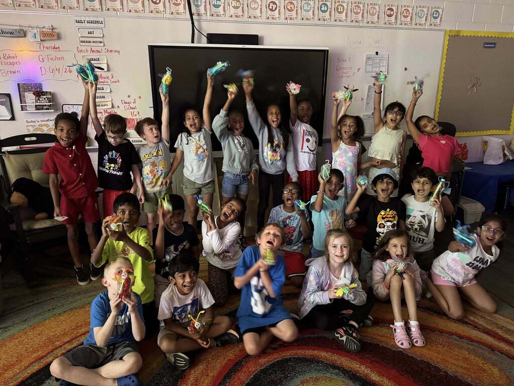 Group of young children in a classroom holding up colorful craft items, smiling and celebrating together on a patterned rug.