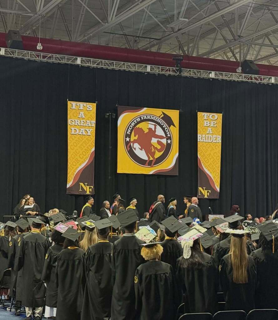 Graduates in black caps and gowns gather in an auditorium beneath banners reading 'It's a great day to be a Raider' and a North Farmington logo.