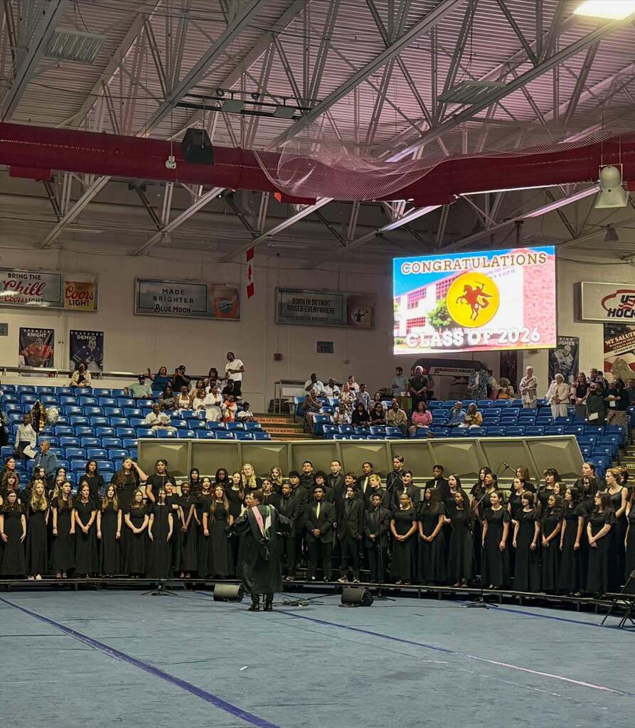 Graduating students in black gowns stand on stage in a gymnasium with a congratulatory sign for the Class of 2026 behind them.