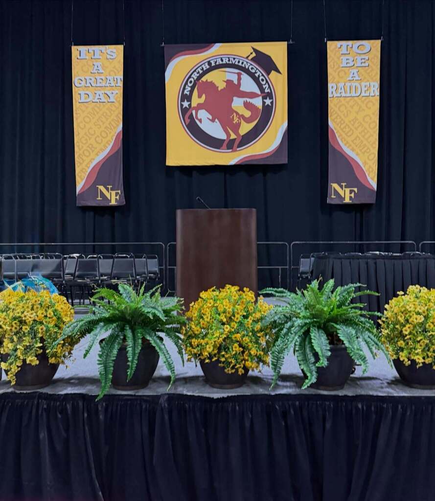 Stage setup with podium and banners displaying North Farmington Raider logo and slogans, decorated with yellow flowers and green ferns.