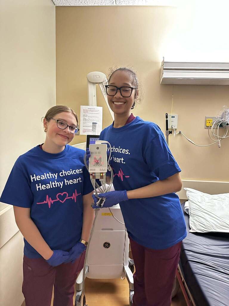 Two healthcare workers in blue shirts with 'Healthy choices. Healthy heart.' stand beside medical equipment in a hospital room.