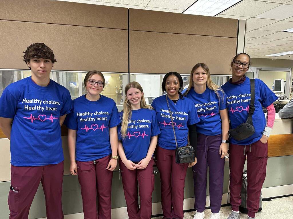 Group of six people wearing matching blue shirts with 'Healthy choices. Healthy heart.' and maroon pants standing indoors.