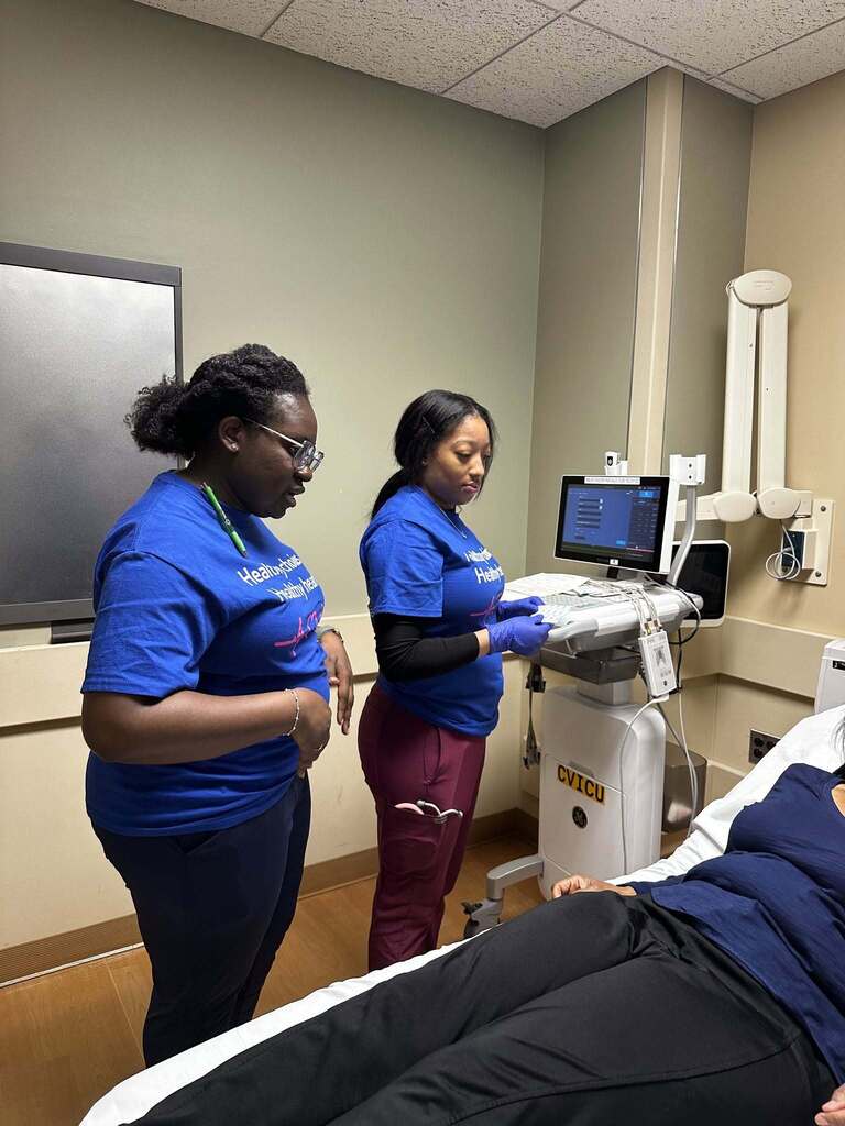 Two healthcare workers in blue shirts operate an ultrasound machine beside a patient lying on a hospital bed in a clinical room.