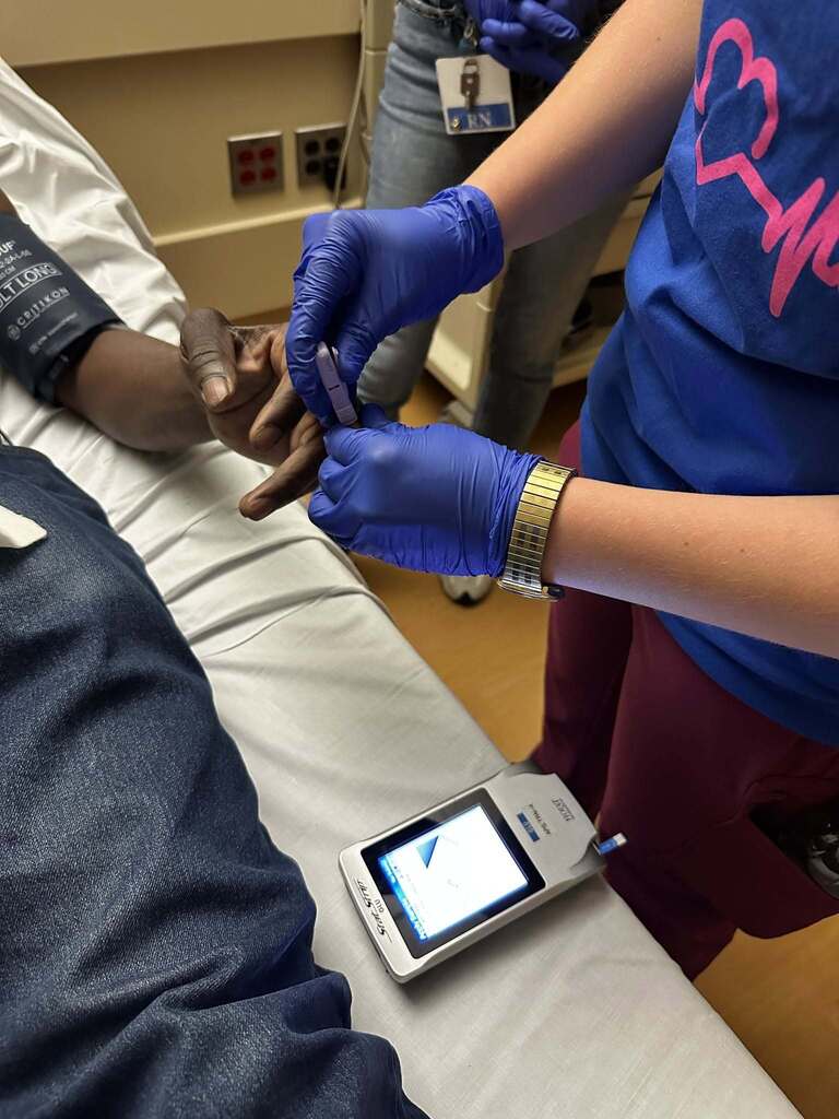 Healthcare worker wearing blue gloves performing a finger prick blood test on a patient lying in a hospital bed.