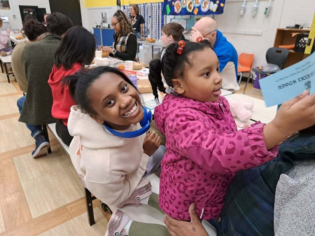 Two students sitting at a table. One is leaning in close to the camera and smiling.