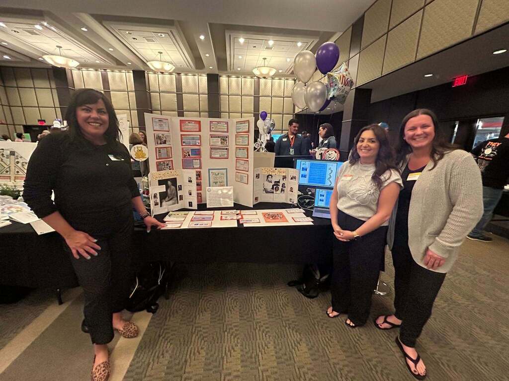 Three smiling women stand next to a conference display