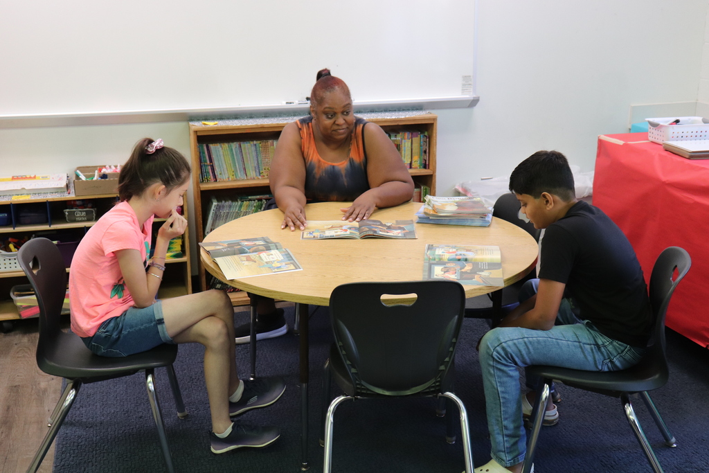 Teacher and two students sitting around a round table reading books in a classroom setting.