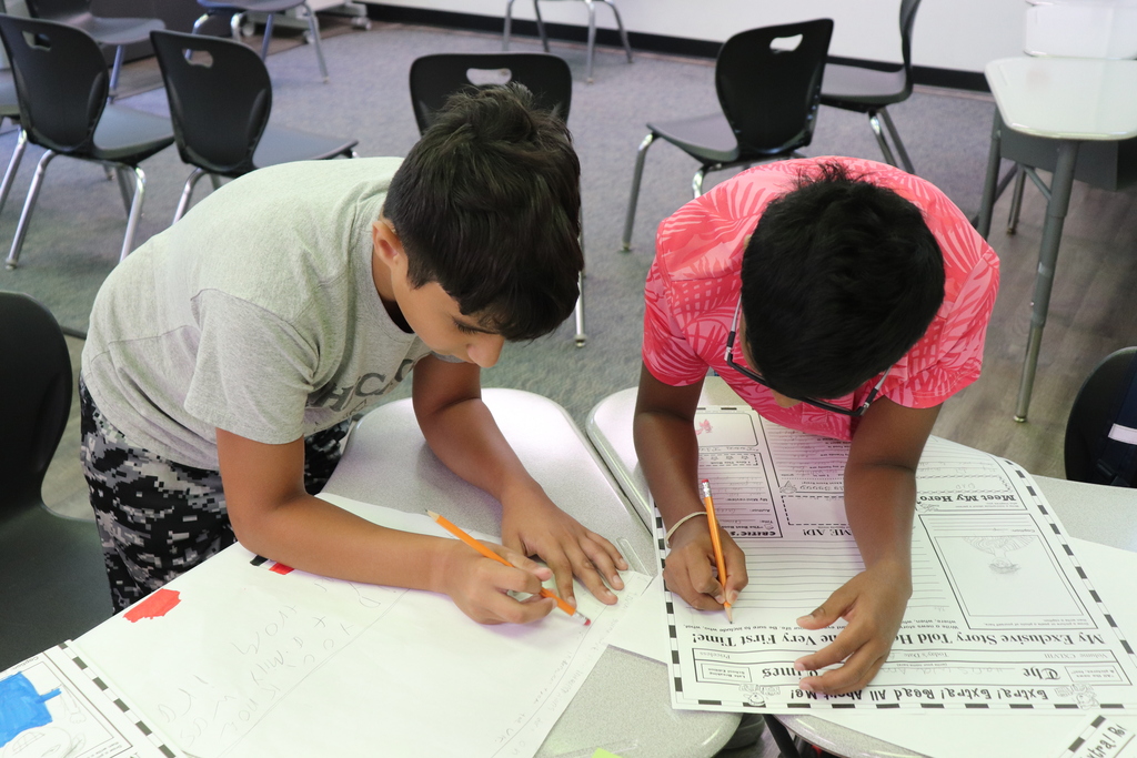 Two boys leaning over a table, focused on writing and drawing on large sheets of paper in a classroom setting.