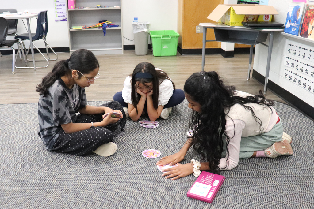 Three girls sitting on a classroom carpet playing a card game together, focused and engaged.