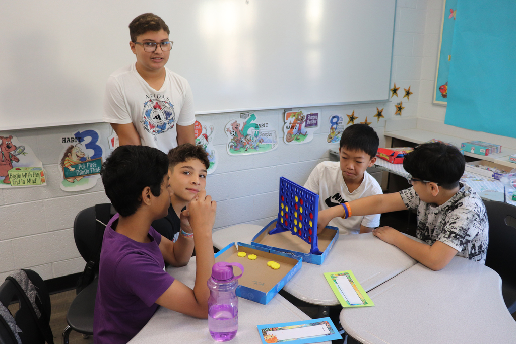 Five boys gathered around a table playing Connect Four in a colorful classroom setting with educational posters on the wall.