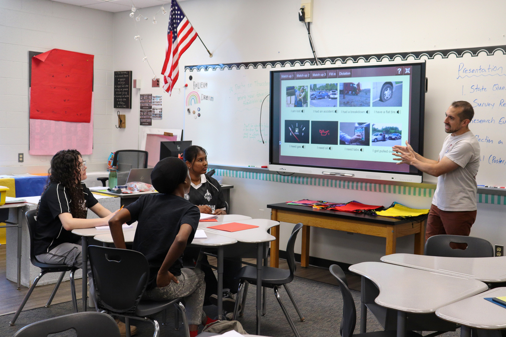 Teacher presenting digital images on a screen to three attentive students seated around a classroom table.