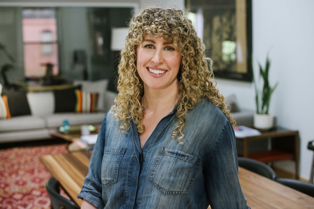 Smiling woman with curly blonde hair wearing a denim shirt standing in a cozy living room with a wooden table and plants.