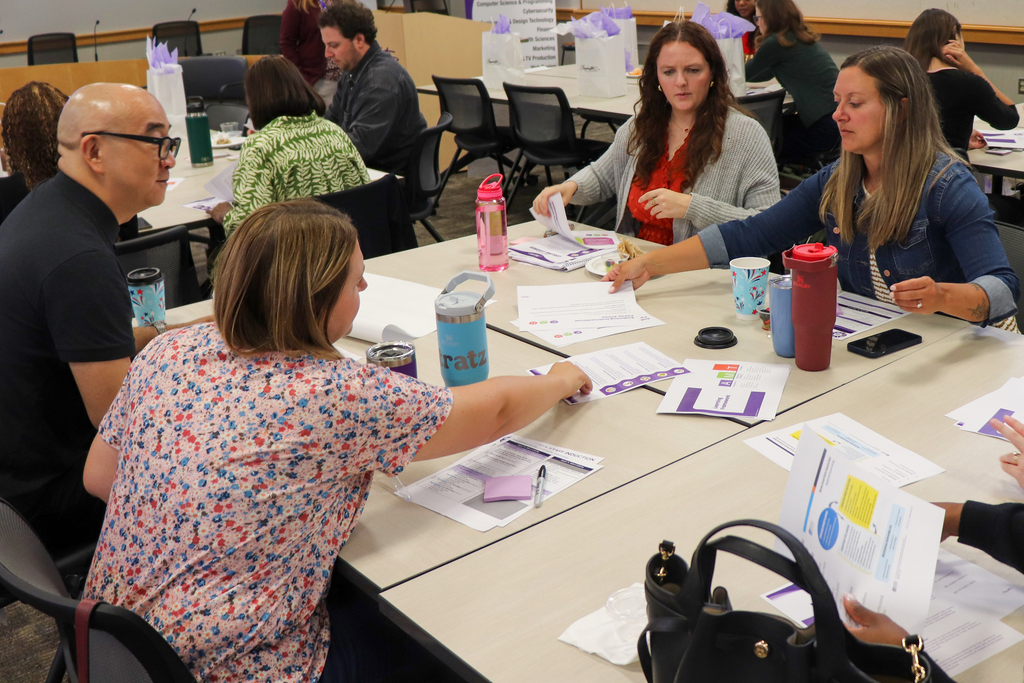 A group of adults engaged in a collaborative meeting around a table filled with papers, water bottles, and coffee cups.