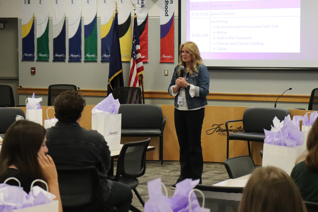 Woman speaking in front of seated audience during a presentation in a conference room with flags and a projection screen.