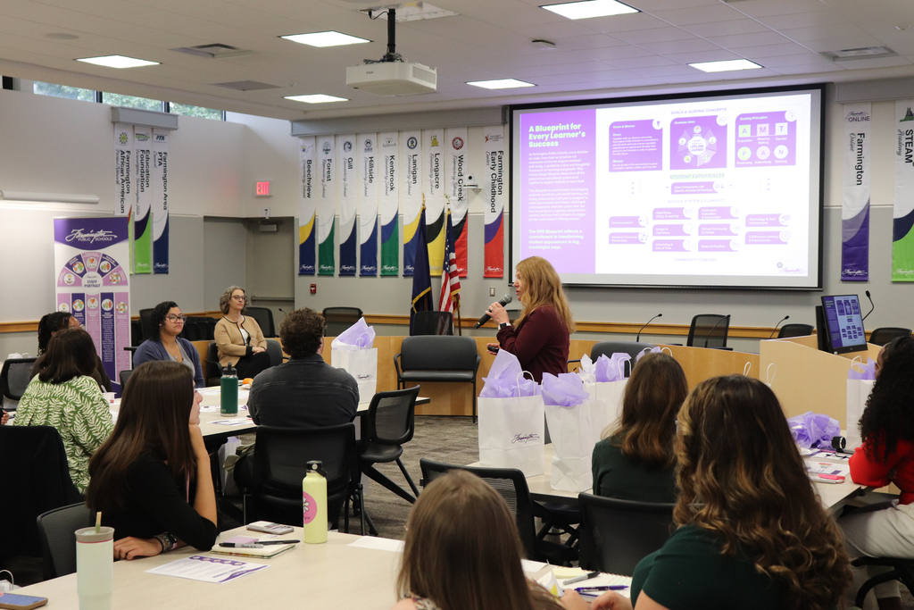 Woman presenting data on a large screen to an audience seated around tables in a conference room decorated with colorful banners.