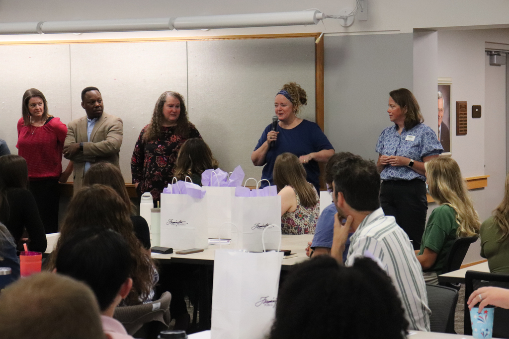 Group of four speakers addressing an attentive audience seated at tables with gift bags in a conference room.