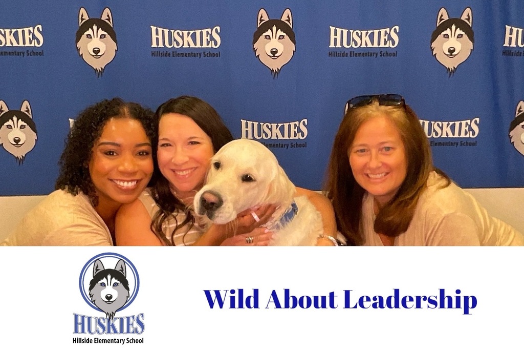Three smiling women pose closely with a white dog against a blue backdrop featuring Huskies logos from Hillside Elementary School.