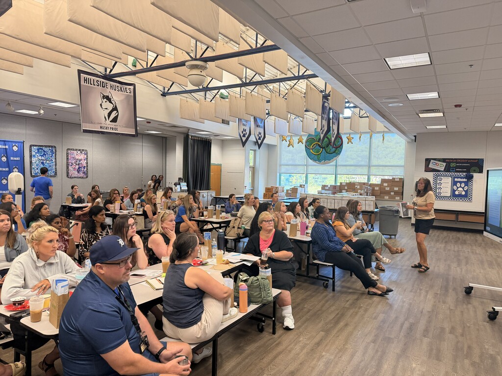 A woman stands addressing a large group seated at tables in a school cafeteria decorated with banners and posters.