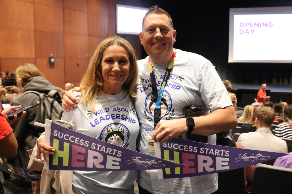 Two smiling adults wearing 'Wild About Leadership' shirts hold a banner reading 'Success Starts Here' at an indoor event.