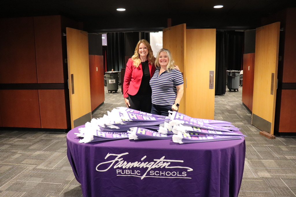 Two women standing behind a table covered with a purple Farmington Public Schools cloth and stacks of folded purple umbrellas.