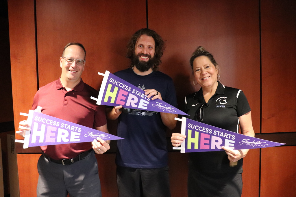Three smiling adults holding purple pennants that read 'Success Starts Here' against a wooden wall background.