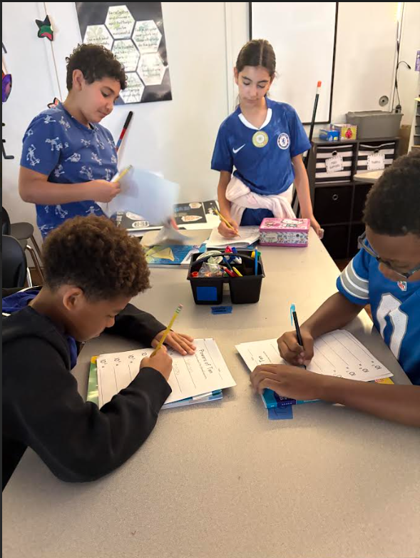 Four children engaged in writing activities at a classroom table with school supplies scattered around.