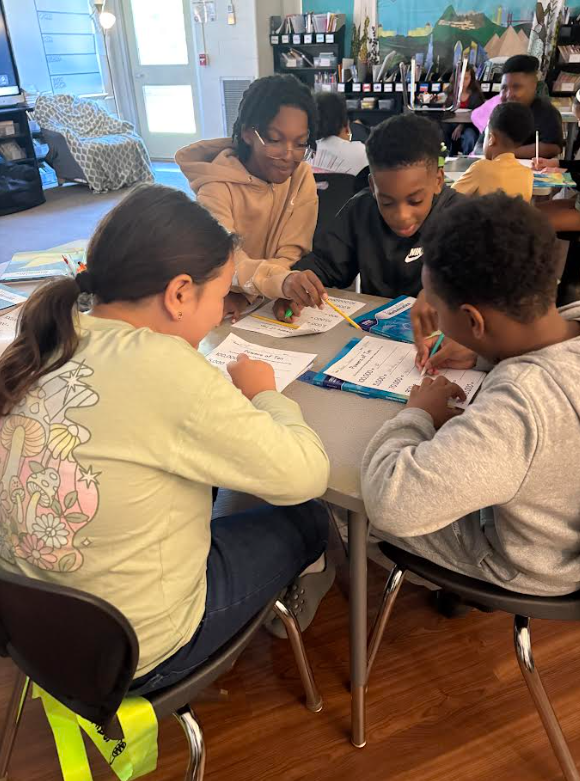 Four students collaborate on a classroom assignment, writing and discussing papers around a round table.