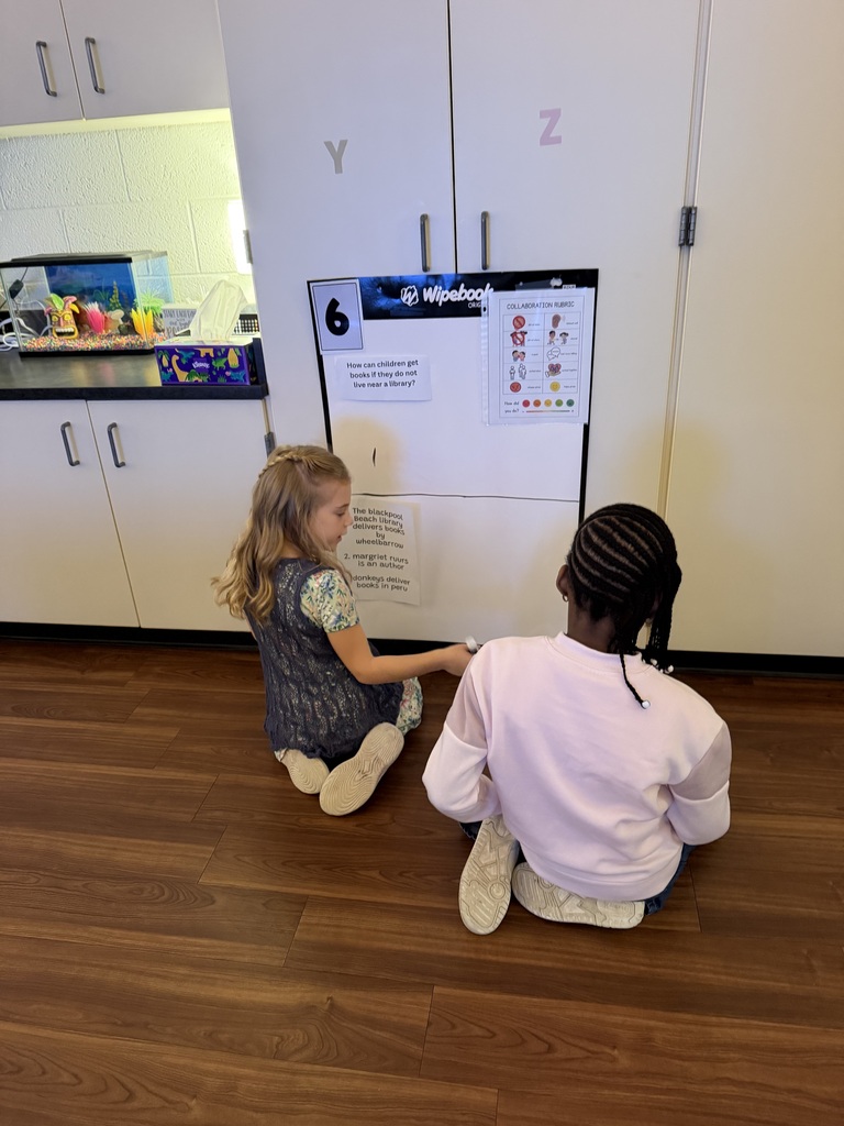 Two young girls sitting on the floor, closely examining a whiteboard with drawings and educational text about how children get ideas for stories.