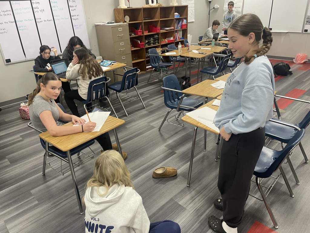 Students in class standing, sitting at desk, and sitting on the floor