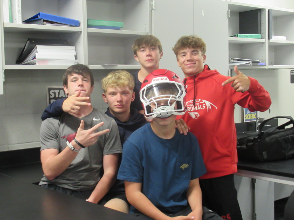 Five boys sitting in class together one with helmet on head.