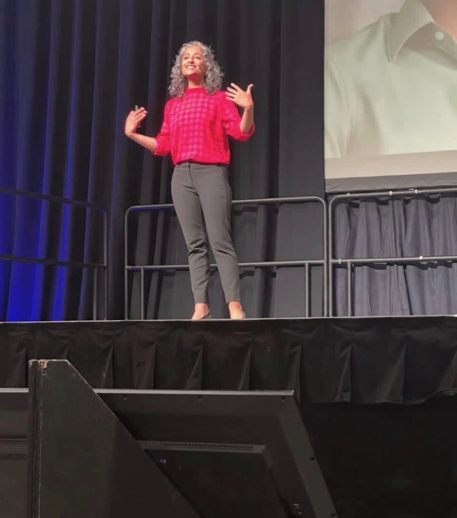 A woman stands on a stage, gesturing with raised hands. Behind her, a screen displays a man in a white shirt. The stage has a blue backdrop and black curtains.