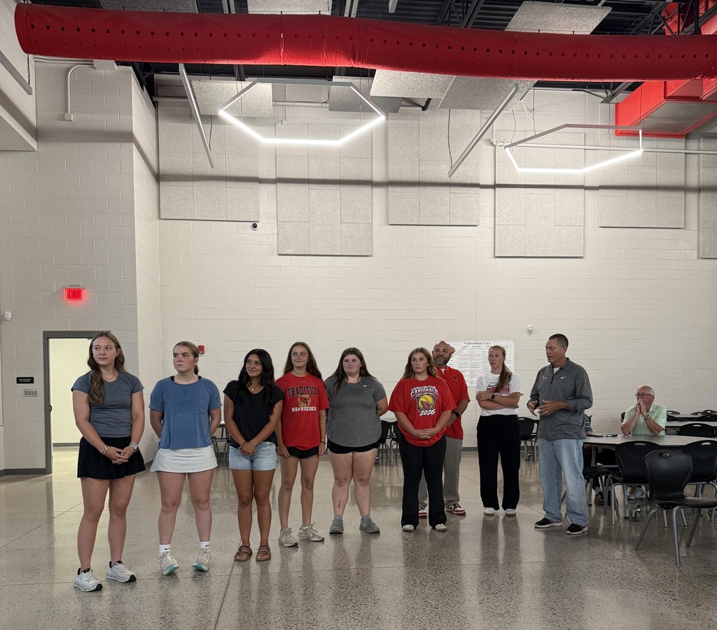 Nine people in a large room stand in two lines. A man stands in front of them. A door is in the left background.