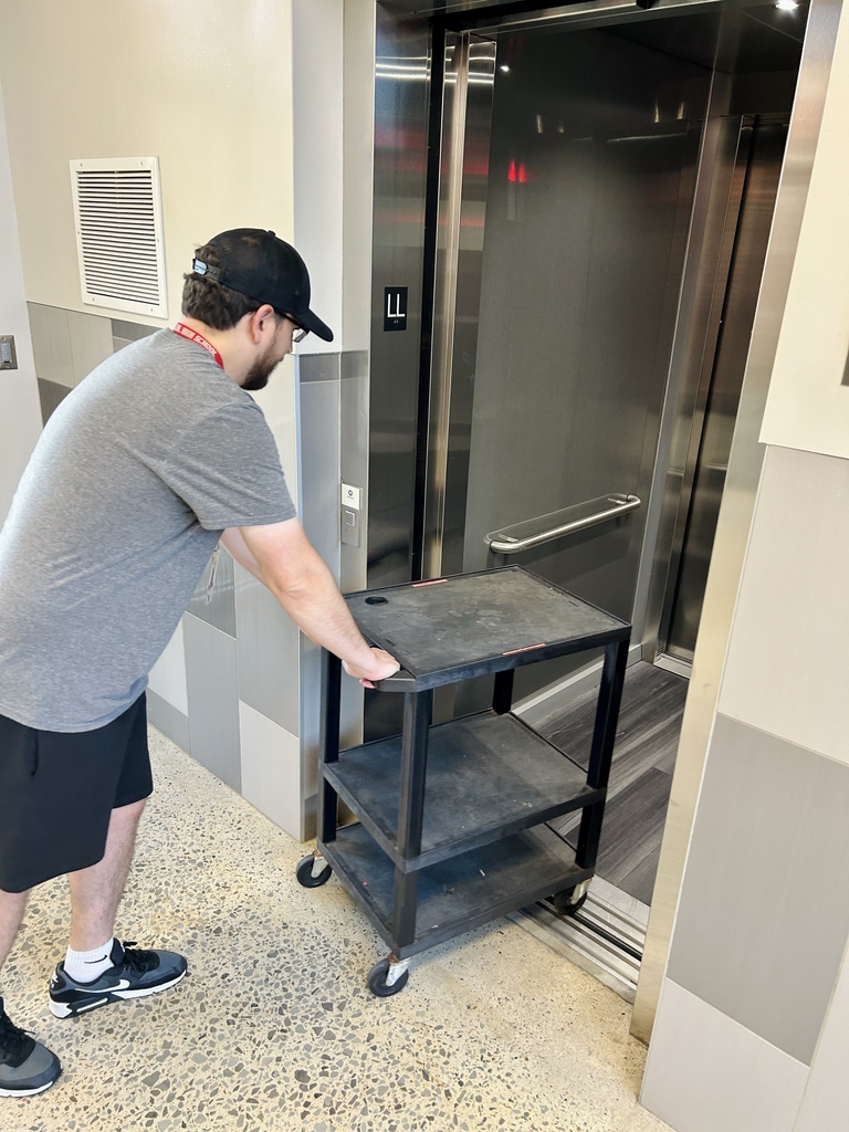 A person wearing a gray shirt, black shorts, and a cap wheels a cart through an open elevator.
