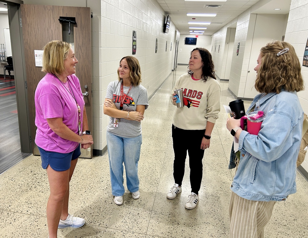 Four women in casual clothes stand in a hallway. One holds a camera, and another carries a book.