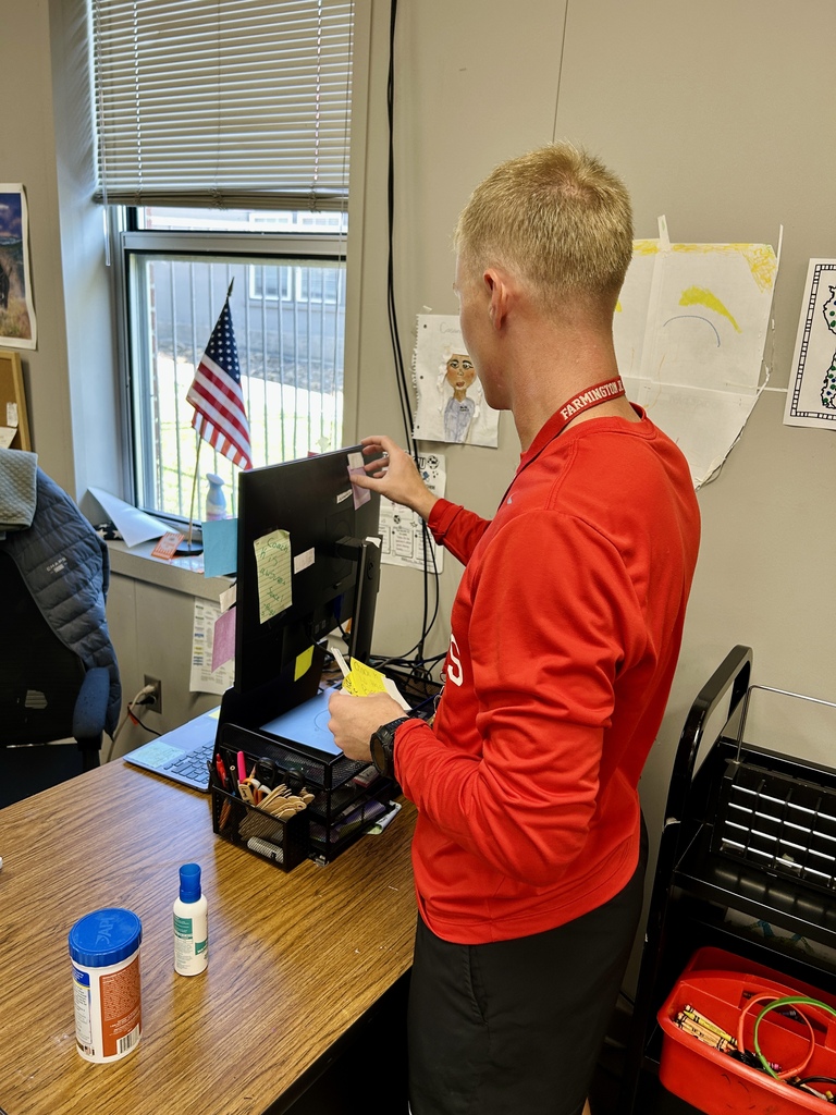 Man in red long-sleeve shirt and black pants working on a device on a table. Behind him, an American flag.