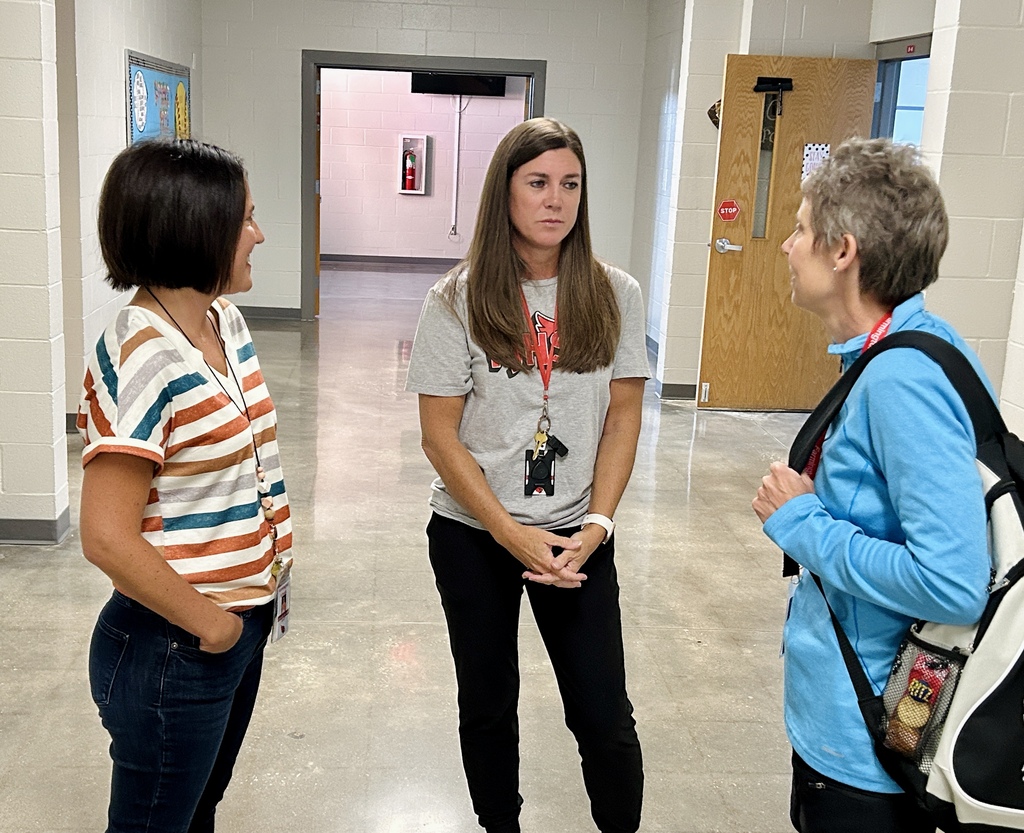 Three women converse in a hallway with white walls and a door. One wears a backpack.