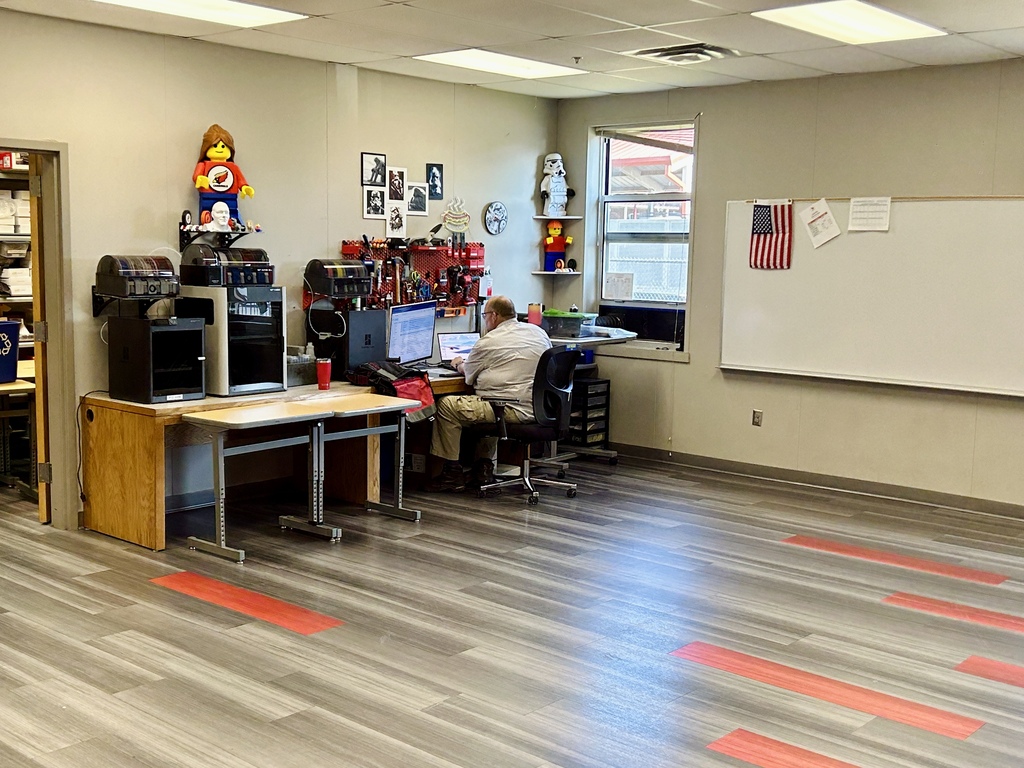 A person at a computer on a desk, with an American flag on the wall, a whiteboard, and a toy in the room.