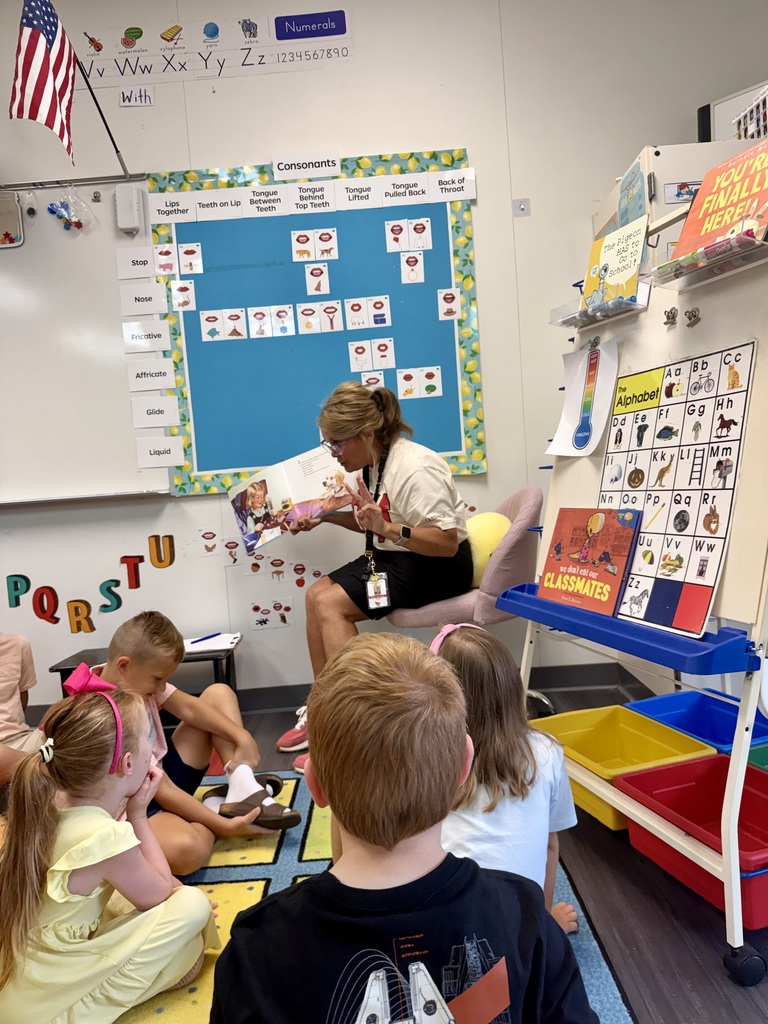Teacher reading to students in a classroom with an alphabet board and colorful bins.