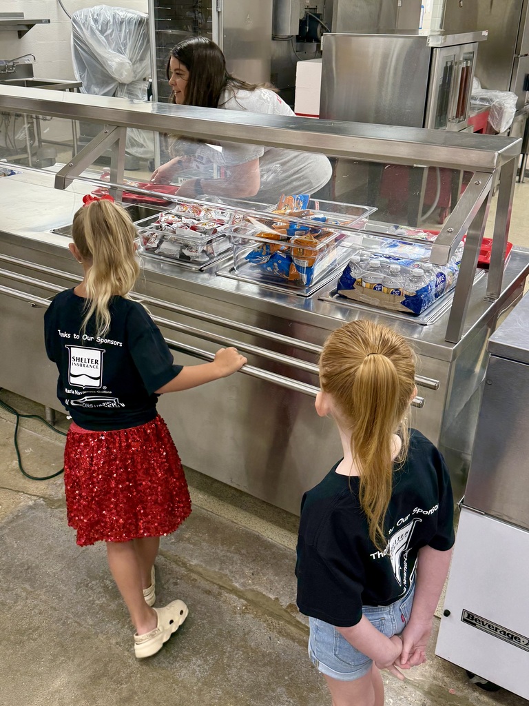 Two young girls in front of a food counter, one holding a shelf edge. A person in a white shirt works behind the counter.