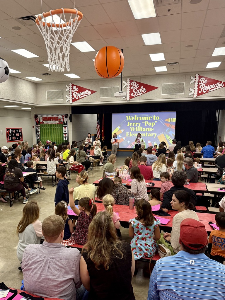 A room with tables and chairs where a large group of people are seated. A basketball hoop is hanging from the ceiling.